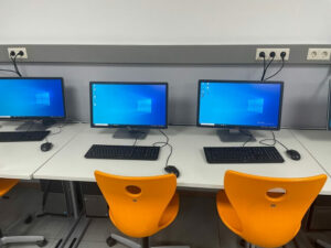 drei Arbeitsplätze mit einem angeschalteten PC, Tastatur und einem orangefarbenen Stuhl